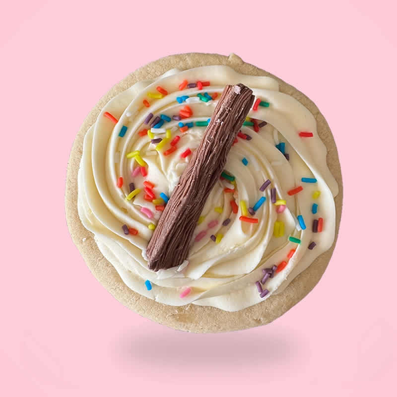 Cookie with white frosting, colorful sprinkles, and a chocolate stick on a pink background